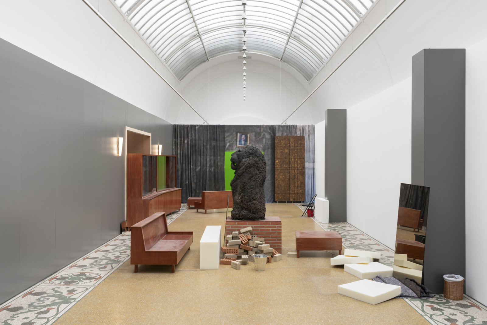 Abbas Akhavan, cast for a folly, 2019/2023, museum vitrines, benches, false doorway, digitally printed sharkstooth scrim fabric wall, green screen, mirrors, sconces, oscillating fan, chairs, plastic bucket, air conditioner, open cell foam cushions, moving blanket, clay bricks, pump, water lilies, dolly, straw, sand, plywood, mortar, dimensions variable. Installation view, curtain call, Ny Carlsberg Glyptotek, Copenhagen, Denmark, 2023
