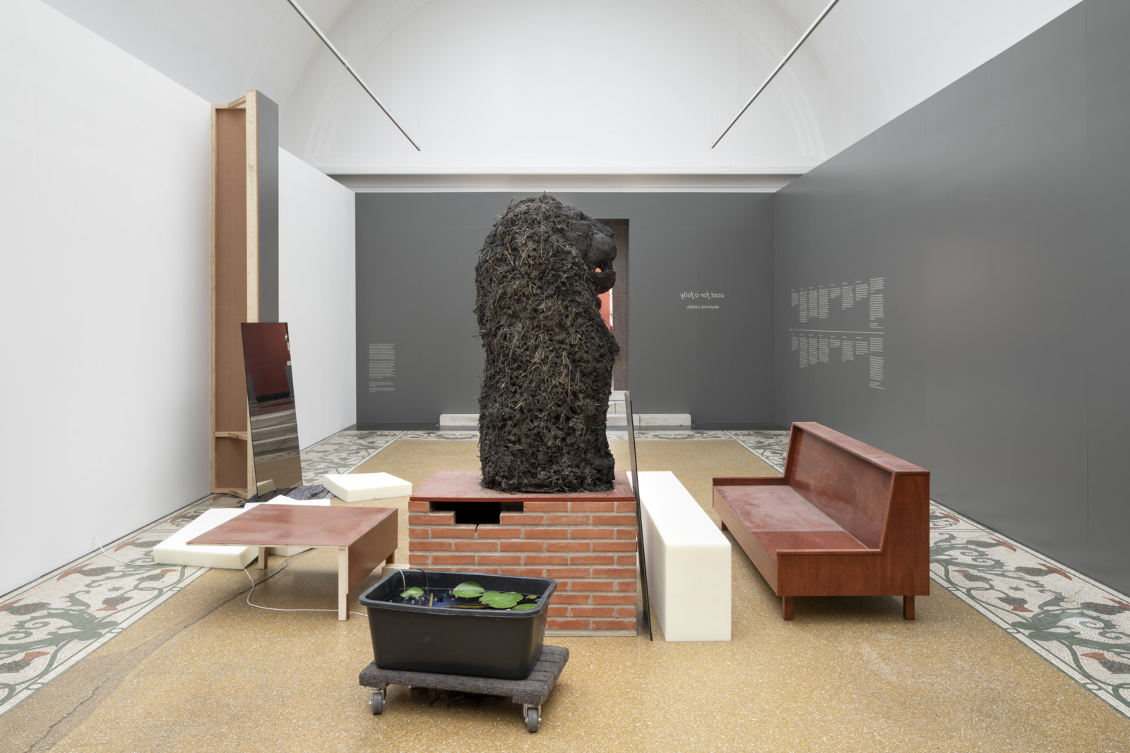 Abbas Akhavan, cast for a folly, 2019/2023, museum vitrines, benches, false doorway, digitally printed sharkstooth scrim fabric wall, green screen, mirrors, sconces, oscillating fan, chairs, plastic bucket, air conditioner, open cell foam cushions, moving blanket, clay bricks, pump, water lilies, dolly, straw, sand, plywood, mortar, dimensions variable. Installation view, curtain call, Ny Carlsberg Glyptotek, Copenhagen, Denmark, 2023