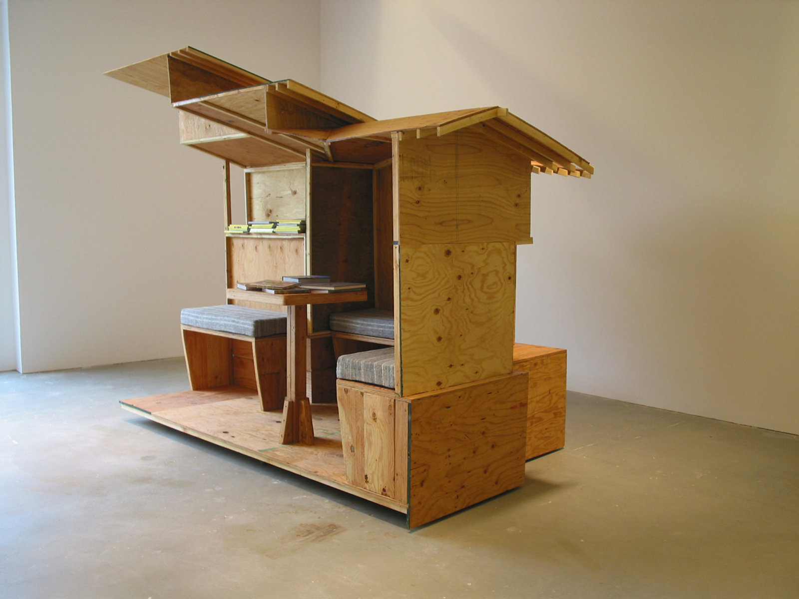 Brian Jungen, Arts and Crafts Book Depository / Capp Street Project 2004, 2004, architectural model (scale of Greene and Greene's Gamble House) made of plywood sectioned into 4 quadrants, locking castors, bookshelves, 2 framed glass cabinets with electrical source and lighting unit, hand-made fabric pillows for seating benches, video monitor, ongoing accumulation of library inventory of magazines, journals, books and videos, 108 x 192 x 252 in. (274 x 488 x 640 cm)