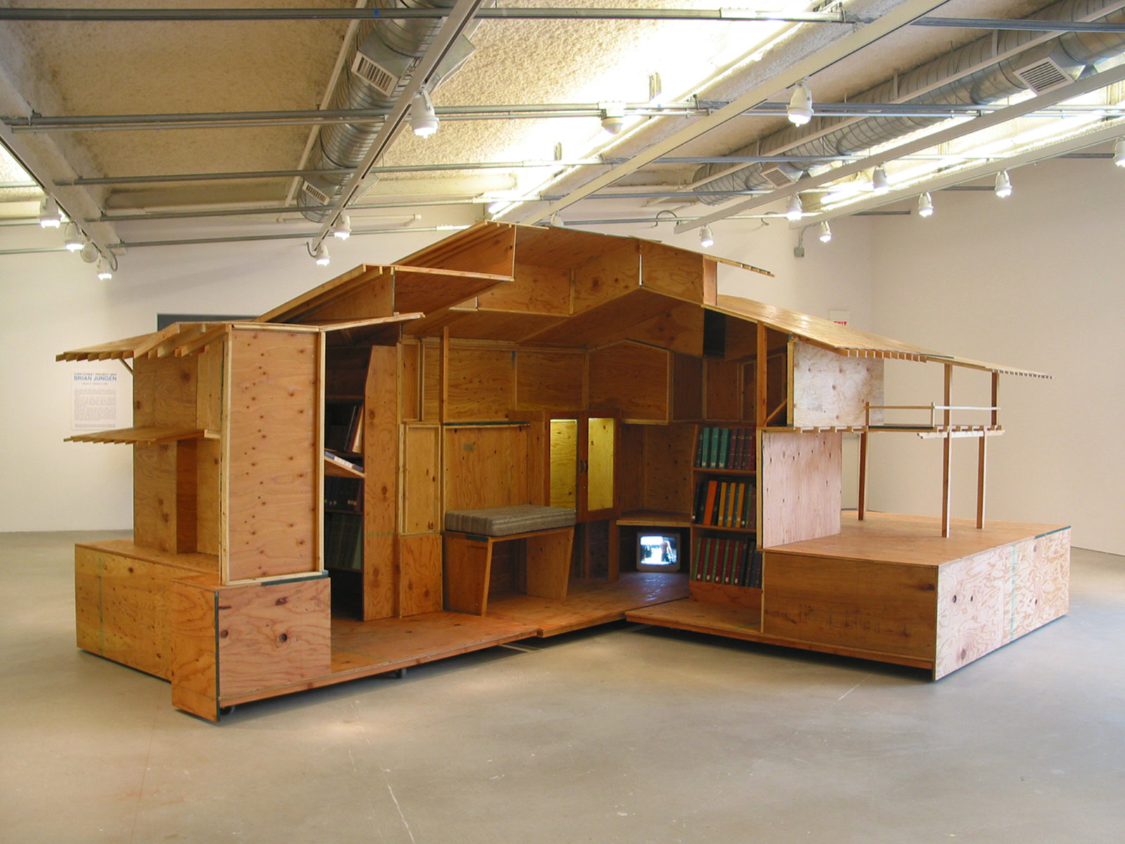 Brian Jungen, Arts and Crafts Book Depository / Capp Street Project 2004, 2004, architectural model (scale of Greene and Greene's Gamble House) made of plywood sectioned into 4 quadrants, locking castors, bookshelves, 2 framed glass cabinets with electrical source and lighting unit, hand-made fabric pillows for seating benches, video monitor, ongoing accumulation of library inventory of magazines, journals, books and videos, 108 x 192 x 252 in. (274 x 488 x 640 cm)