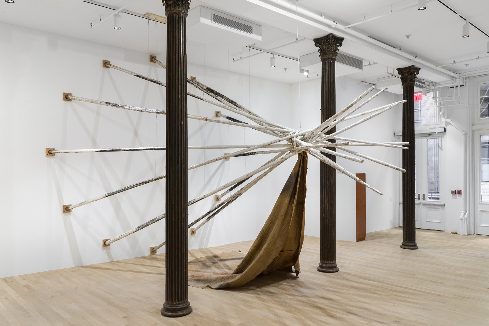 Duane Linklater, dislodgevanishskinground, 2019, 12 teepee poles, steel cable, white paint, charcoal, rope, digital print on linen, black tea, blueberry extract, sumac, charcoal, 220 x 174 x 174 in. (559 x 442 x 442 cm). Installation view, Artists Space, New York, USA, 2019