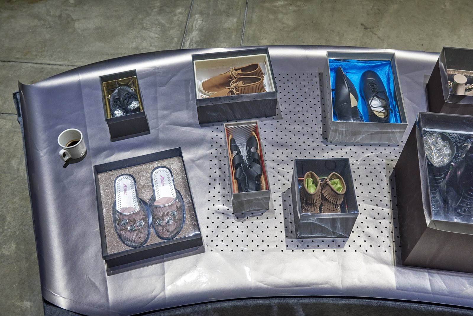 Liz Magor, Shoe World (detail), 2018, 33 pairs of secondhand shoes, mat-board shoeboxes of variable dimensions with polyester film lids, porcelain mug, coffee, and plywood platform with fabric cover, 12 x 288 x 40 in. (31 x 732 x 102 cm). Installation view, BLOWOUT, Carpenter Center for Visual Arts, Cambridge, USA