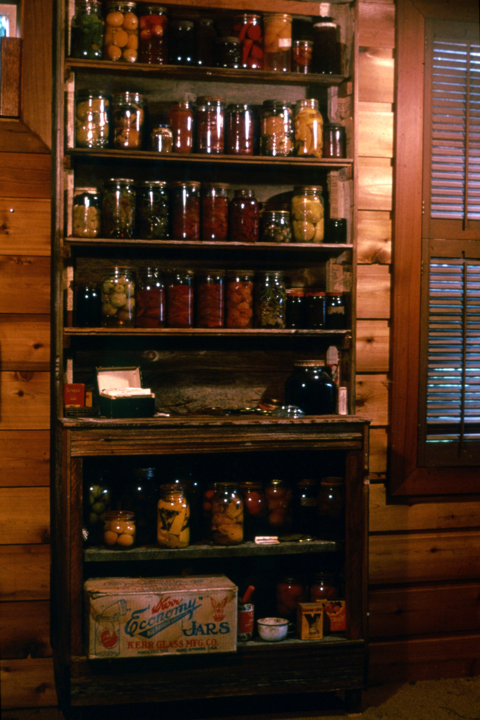 Liz Magor, Time and Mrs. Tiber, 1976, wooden shelf with jars of preserves, recipe box, forks, glass tops, rubber sealers, metal lids, cardboard boxes, enamel cup, tin can, 84 x 36 x 13 in. (214 x 91 x 32 cm)
