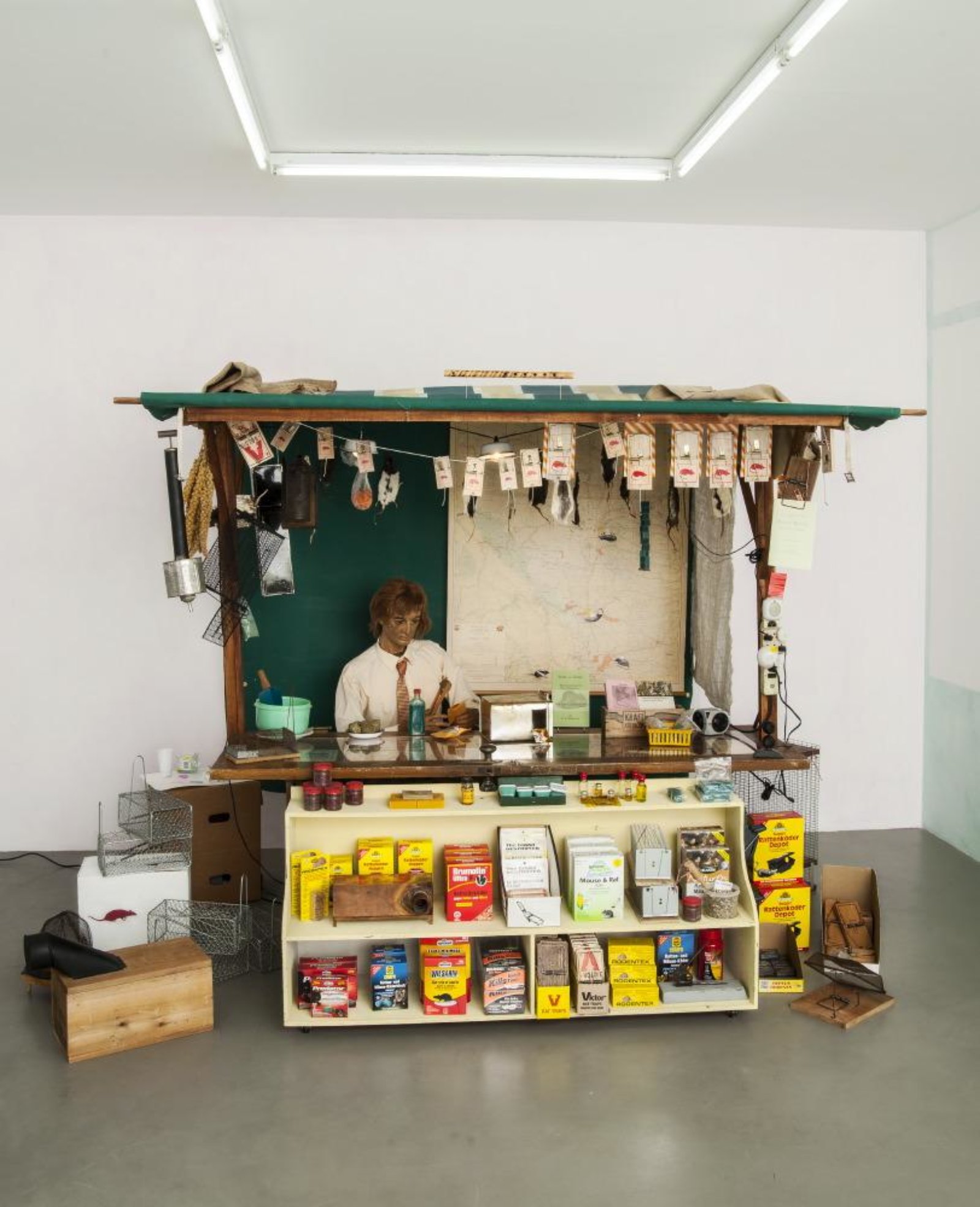 Ashes Withyman, An Ultrasonic Flute, 2013–2014, main structure: wooden framework, painted canvas, map of Alberta with collage, press-on letter-sign, folded metal wheeled carrier, folding steel chair, wax mannequin, pencil crayon drawing of rat, sign (collage and paint on cardboard), burlap sack, piece of linen, home-made smoker, blue ribbon millet, rat bait, rat and mouse traps, rat food, rat pelts, Chinese steel rat cages, book page, pesticide sprayer, leather and wood bellows, string lighting with pie plates, rat food on tin plate, envelope with label, black rat trap, rat poison bottle, rat tail cutter, kraft single cheese box, two ultrasonic deterrents, burrow smoker, 3 ultrasonic devices attached to power bar, cardboard box, wood and metal rat trap, tomato tin rat trap, folding table made from door with plexiglas top; under plexiglas top: magazine clipping, extermination booklet, vintage label, dream almanac, white rat pelt, 5 zip lock food bags with cartoon mouse label, 3 booklets; on table top: rat food in tupperware container with home-made scoop, rat bait in zip lock bags in green tin, circular rat trap, glass bottle with rat and blue liquid, rat food in tupperware with hand carved wooden spoon; plywood display shelf: ziploc bag of mock rat bait, herbal oils in yellow plastic basket, rubber gloves with business cards, 4 tupperware containers of green rice, old rat trap, two home-made traps, box of “The giant destroyer” rat catchers, “Nooski” rat traps, “Green Earth” mouse and rat bait, “Wilsarin” rat bait, “Predator” rat bait, tupperware containers of red rice, viktor rat traps, rat trap boxes, 2 home-made rat traps, “Rodentex” mouse bait; dimensions variable